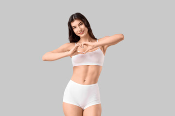 Fototapeta premium Pretty young woman in underwear making heart with her hands on light background