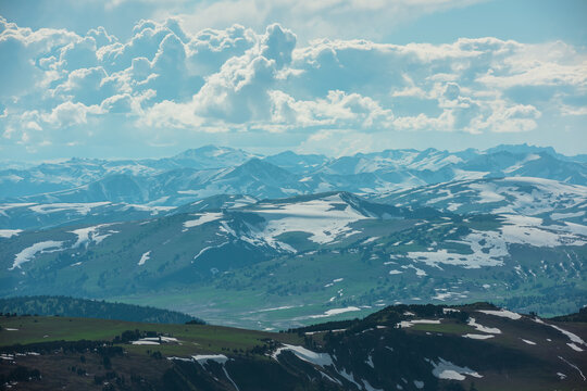 Aerial sunlit top view above forest hills and big snowy mountain range under lush clouds in changeable weather. Scenic hilly alpine landscape with sunlight and shadows. Partly cloudy in high mountains