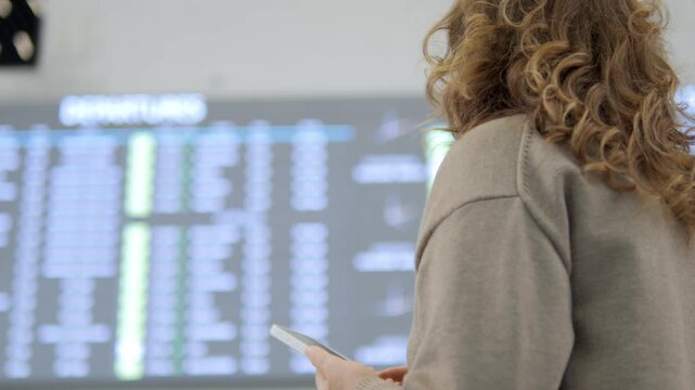 Woman with curly hair stands near airport departure board. She holds phone, looks at flight information. Her posture shows patience, slight worry about delay. Suitable for travel, tourism