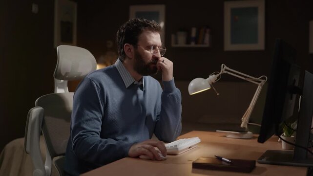 Freelancer thinking, pensive man. Male freelancer stares thoughtfully at computer screen, occasionally typing on keyboard. Camera slowly zooms in