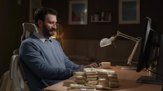 Success, high-paying job, high income concept. Male freelancer, remote worker is typing at computer with huge pile of cash lying on desk nearby.