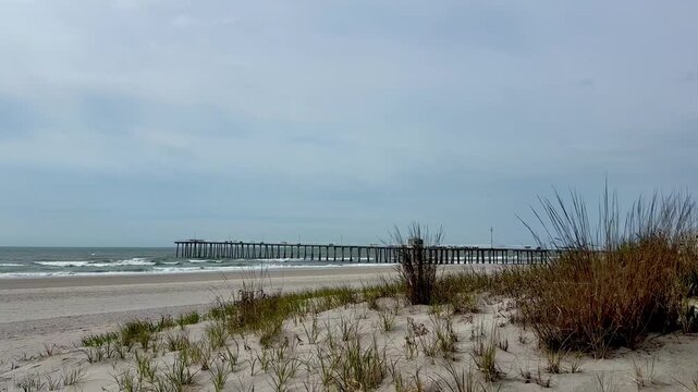 Descending view from blue sky down to ocean, pier and grassy sand dune.