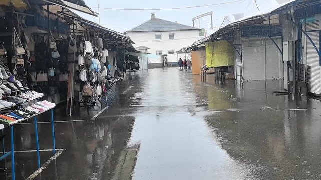 Rain pours on a quiet market in the city with closed shops and water collecting on the ground. Few people walk through as they avoid the wet conditions.