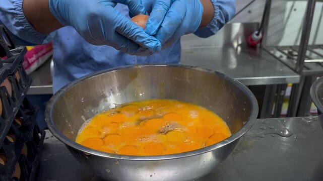 Worker cracking eggs into a mixing bowl during food manufacturing operations. Ingredient preparation process supporting food production, quality control, and hygiene standards in a food processing env