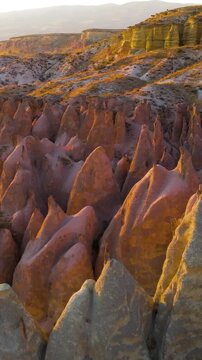 Vertical video. Goreme, Nevsehir, Turkey. Devrent Valley fairy chimneys, low altitude aerial flight over whimsical eroded rock spires glowing in warm golden hour sunset light. Aerial View. Rich color