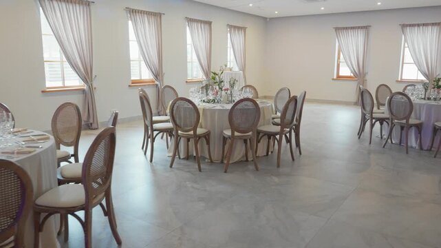 Bright, airy event space with round tables dressed in white linens, elegant settings, fresh flowers, and soft natural light. Wide-angle pan reveals tastefully decorated venue, no people, modern minima