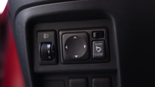 Close-up of a car's interior control panel. Visible are the side mirror adjustment buttons, auto-close switch, and headlight leveling dial on a textured black dashboard.