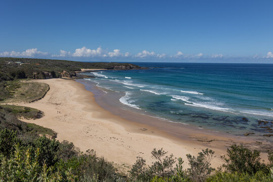 A wide sandy beach with waves in a gorgeously colored sea showing rocks underwater, headlands with wave cut platforms and a cloudbank in the distance, near Ulladulla, in New South Wales, Australia.