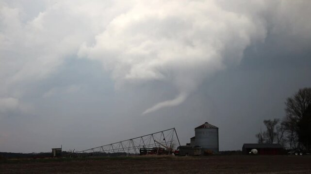 A rope tornado dangles beneath a storm cloud as it passes behind farm buildings on the prairie. Time lapse footage.