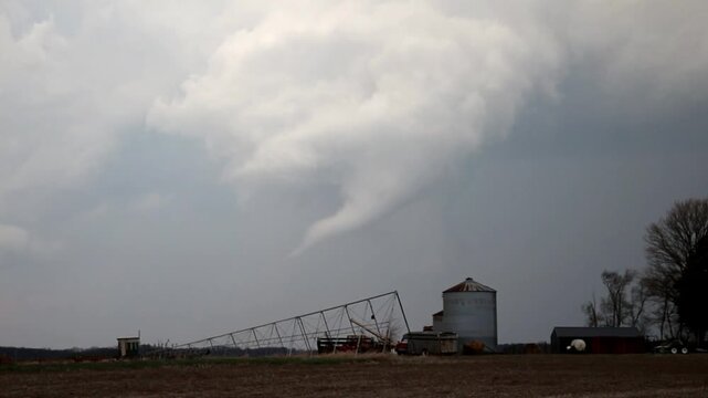 A white tornado drifts through the fields behind a farm. Grain bins and irrigation equipment are seen in the foreground. 