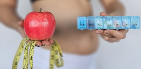 Young plump woman with bare belly stands isolated on white wall holding tape measure apple and diet pills © Nadzeya