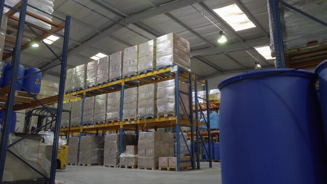 Pan right under soft diffused indoor light across pallet racks, drums and forklift in a coolant fluid warehouse, with an orderly industrial mood.