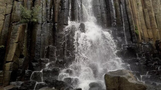 Close view of powerful waterfall cascading over dramatic basalt rock columns with splashing water and rugged volcanic stone texture in Mexico