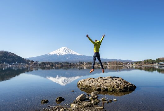 Young man standing on a rock in water, jumping into the air, volcano Mt. Fuji is reflected in Lake Kawaguchi, Yamanashi Prefecture, Japan