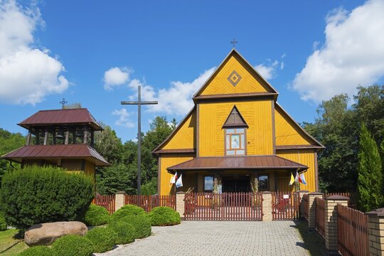 Yellow wooden church in summer under a blue sky with a wooden fence and surrounded by trees, Church of the Holy Apostles Peter and Paul, Olbięcin, Olbiecin, Powiat Kraśnik, Krasnik, Lublin Voivodeship, Poland