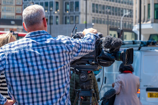 Film crew camera operator tripod street production media event equipment cables van London urban location broadcasting journalism team outdoor coverage setup, Behind-the-scenes television journalism
