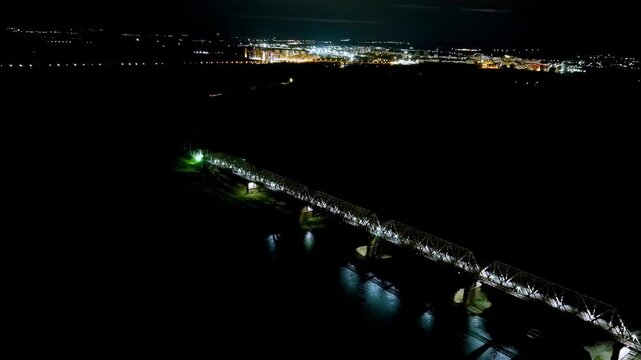 Ufa, Russia. Historic steel railway bridge spanning the Belaya River, its illuminated trusses reflected in dark water against the glowing night cityscape, Drone footage, Departure of the camera