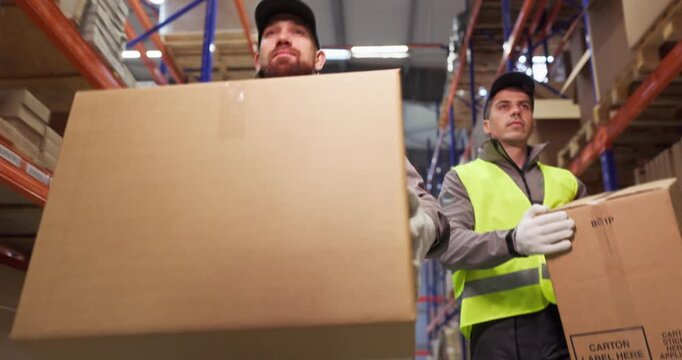 Warehouse workers carrying boxes for delivery. Men in high visibility vests carry cartons through racks, preparing shipping and managing inventory. Clear concept of teamwork and logistics.
