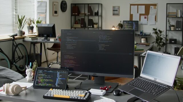 No people medium arc shot of desk of backend software developer at innovative tech startup in office, with laptop, computer and tablet displaying lines of code during script testing or debugging