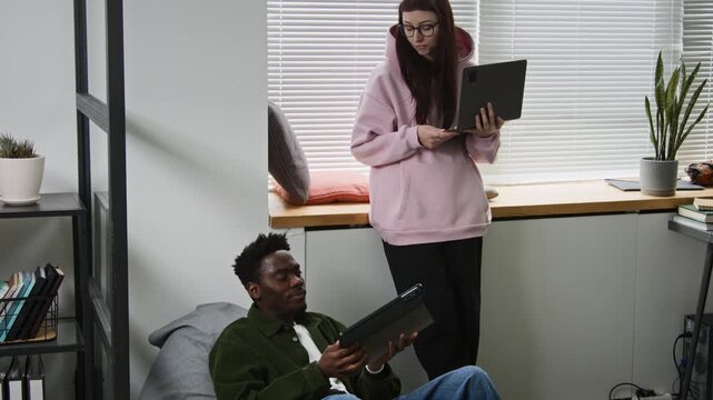 Medium full shot of diverse web developers discussing project in startup office, Caucasian woman with laptop pointing out mistake in code to black male colleague sitting in bean bag chair with tablet