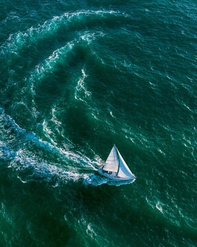 Focused medium drone perspective on a sailing vessel tracing a curved route through intricate emerald wave patterns with the expansive coastal bay gently out of focus emphasizing
