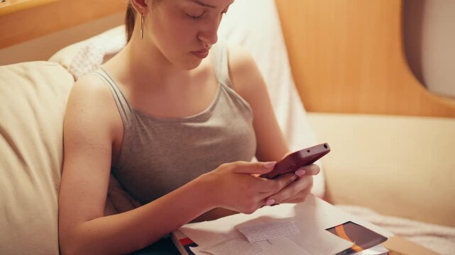 Focused high school girl sitting on a couch and typing on her mobile phone, using a smartphone to copy notes, browse exam answers, or cheat on a future test while studying at home.