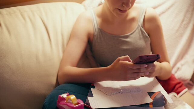 Focused high school girl sitting on a couch and typing on her mobile phone, using a smartphone to copy notes, browse exam answers, or cheat on a future test while studying at home.