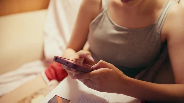 Focused high school girl sitting on a couch and typing on her mobile phone, using a smartphone to copy notes, browse exam answers, or cheat on a future test while studying at home.