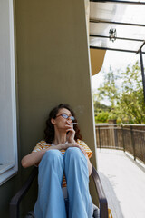 Obraz premium Relaxed woman on a balcony smoking a cigarette, seated in a casual striped shirt and blue pants. Outdoor light creates a calm mood, home lifestyle and quiet contemplation.