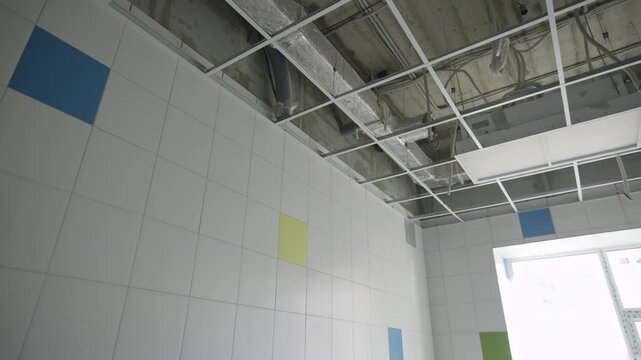 Ceiling grid reveals exposed ventilation pipes above tiled walls. Bright sunshine streams through large square window into empty room