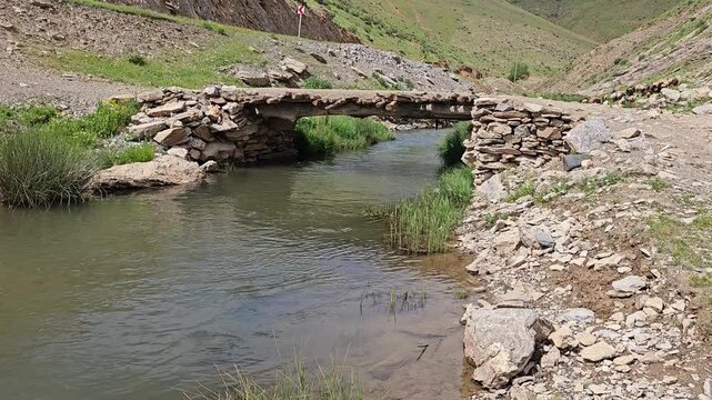 Rocky valley with a flowing stream, an old wooden‑and‑stone bridge, green hills, and wild grasses under a partly cloudy blue sky.
📍 Marivan, Kurdisatn, Iran 