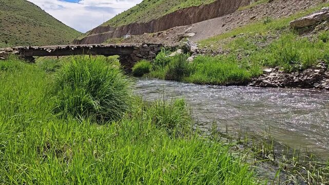 Rocky valley with a flowing stream, an old wooden‑and‑stone bridge, green hills, and wild grasses under a partly cloudy blue sky.
📍 Marivan, Kurdisatn, Iran 