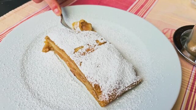 Fork breaks into Austrian apple strudel dusted with powdered sugar on a white plate, flaky pastry with sweet apple filling at an outdoor European cafe restaurant table, dessert close up.