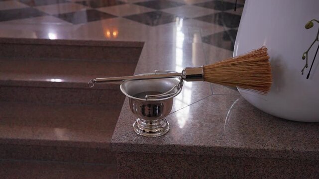 Detailed close-up of a holy water sprinkler and silver bowl resting on polished marble steps in a church. Ambient natural light reflects on the elegant surfaces, evoking a tranquil, spiritual mood.