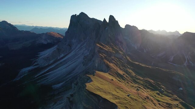 Breathtaking high-angle drone shot of the dramatic Seceda peak and sloped alpine meadows in Val Gardena, Italy. Features majestic jagged mountain ridges illuminated by warm golden hour sunset light.