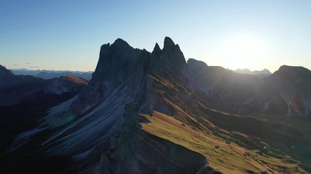 Breathtaking high-angle drone shot of the dramatic Seceda peak and sloped alpine meadows in Val Gardena, Italy. Features majestic jagged mountain ridges illuminated by warm golden hour sunset light.