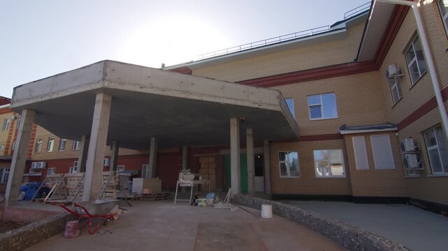 Construction materials lie near new brick building entrance. Sun flare adds warm industrious feel. Unfinished porch attaches to building