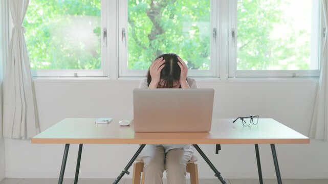 Asian woman holding her head looking distressed while working on laptop