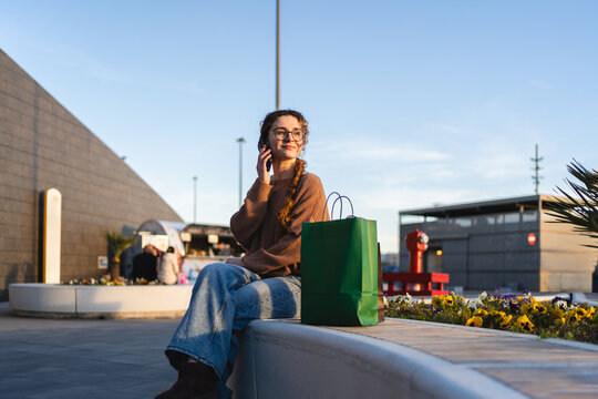 Young woman sitting with shopping bags while using a smartphone at an outdoor mall