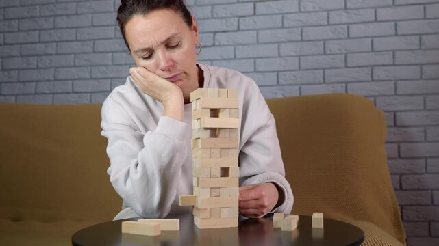 Sad woman playing block stacking board game alone. Depressed woman with a sad expression sitting alone at a table playing a wooden block tower game, showing signs of boredom and loneliness