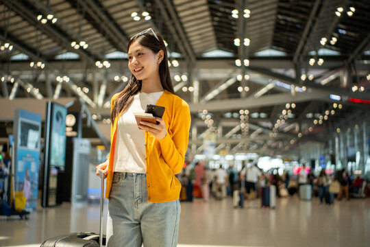 Young asian woman traveler using smartphone for checking flight information check the routes trip while waiting with her luggage at the airport terminal. Traveller woman passport and baggage luggage