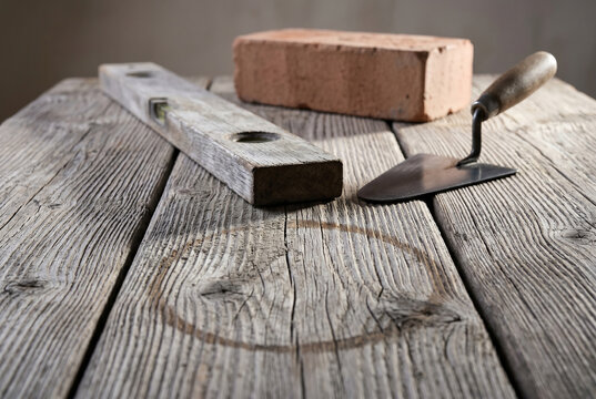 Low angle shot of a time-silvered oak spirit level and a steel trowel on a weathered pine workbench with raised grain and a circular ring stain, under a high-contrast raking light.