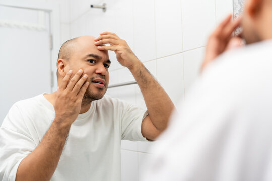 Unhappy young man looking in mirror during morning skincare routine at home. Beauty concerns stress acne problem sensitive skin and self care concept.