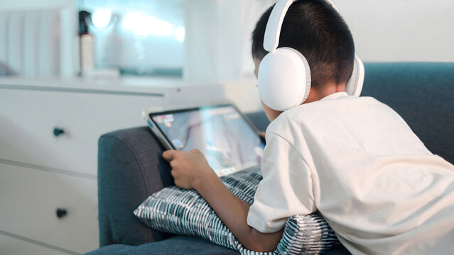 Young boy wearing wireless headphones while using a tablet on a sofa at home. Concept of digital learning, online entertainment, screen time, technology use, childhood lifestyle, and modern parenting.