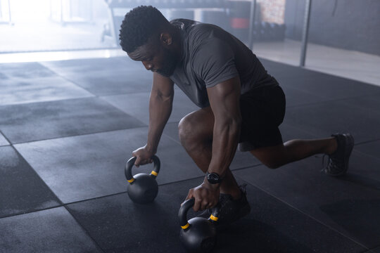 African American man performing kettlebell lunge on gym floor, gripping kettlebells with smartwatch