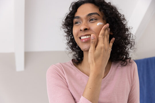 Non-binary person applying white cream to cheek at vanity, wearing pink top, rings and necklace