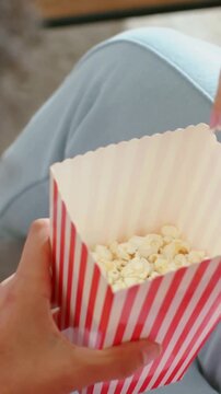 Vertical video: Hovering hand taking popcorn to snack from holder on couch, striped box, pink top