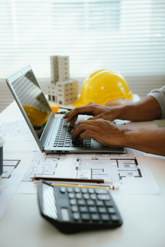 An architect or engineer works on a laptop with building plans and safety equipment, representing construction planning, project management, architecture, and engineering design.
