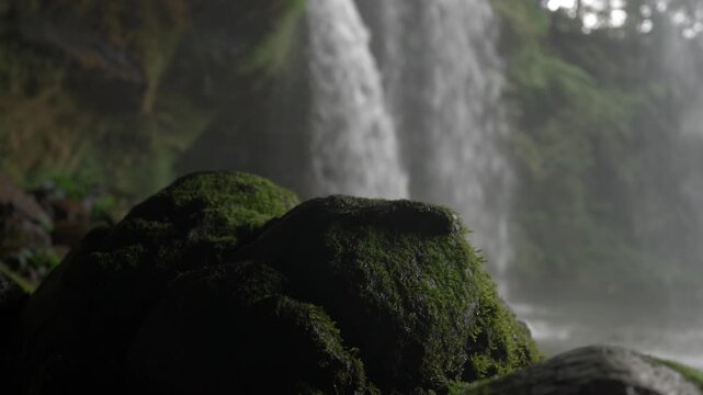 Close up of moss covered rocks beside Tad Champee waterfall, with falling water and mist blurred in the background.
