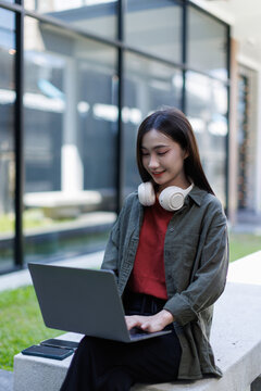 Asian woman student studying online outdoors, using laptop and headphones, engaged in remote learning, distant education, and flexible working from a modern campus